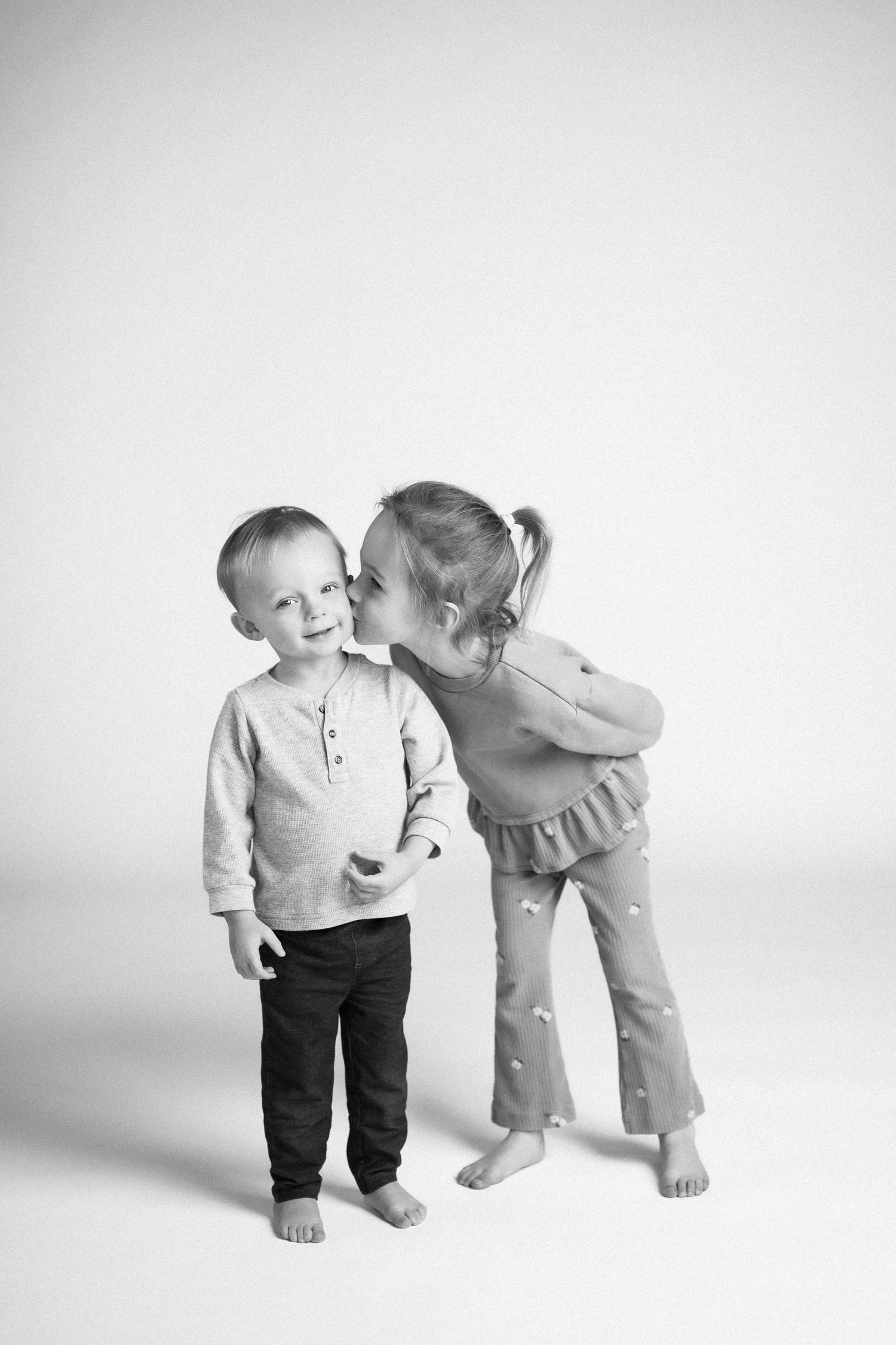 Two young children in casual clothes share a sweet candid moment against a white backdrop in black and white.