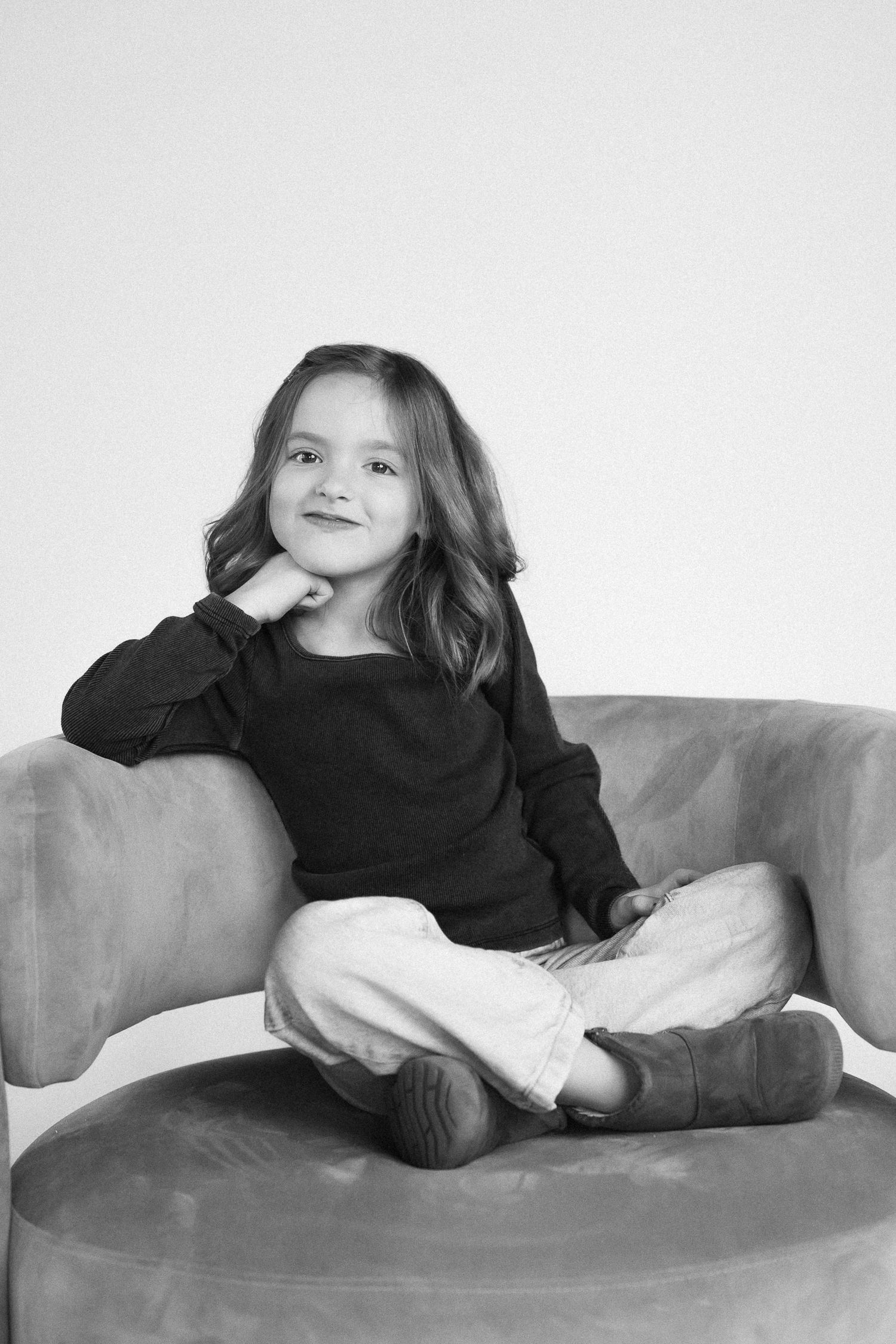 A child in a black sweater and white pants sits cross-legged on a gray couch in a black and white portrait.