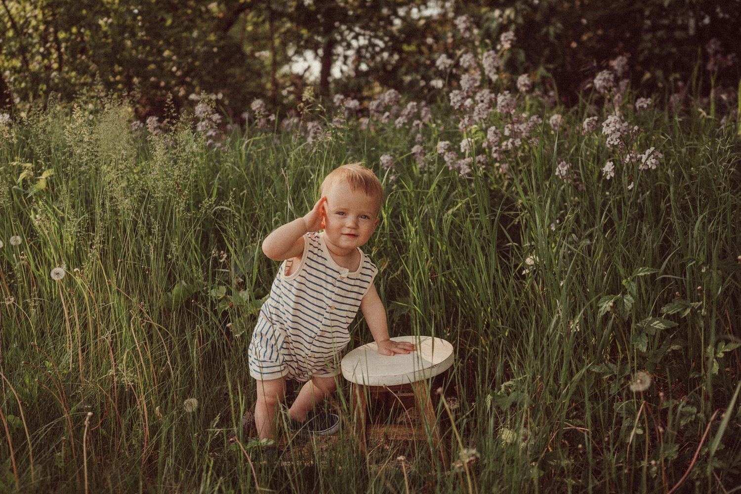 Child in striped clothing crouches by a wooden stump in a wildflower meadow.
