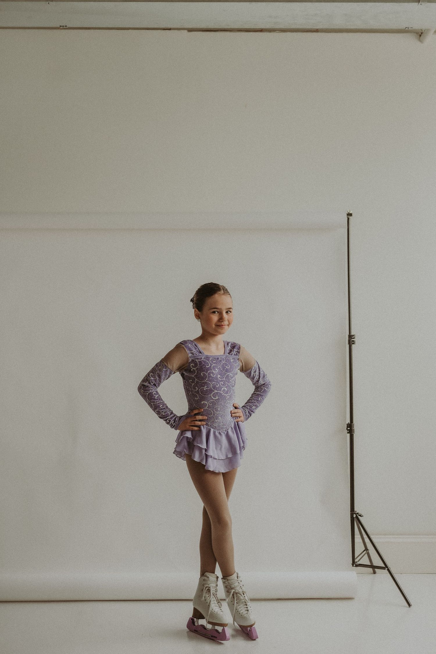 Ice skater stands in purple costume with white skates against plain studio background.