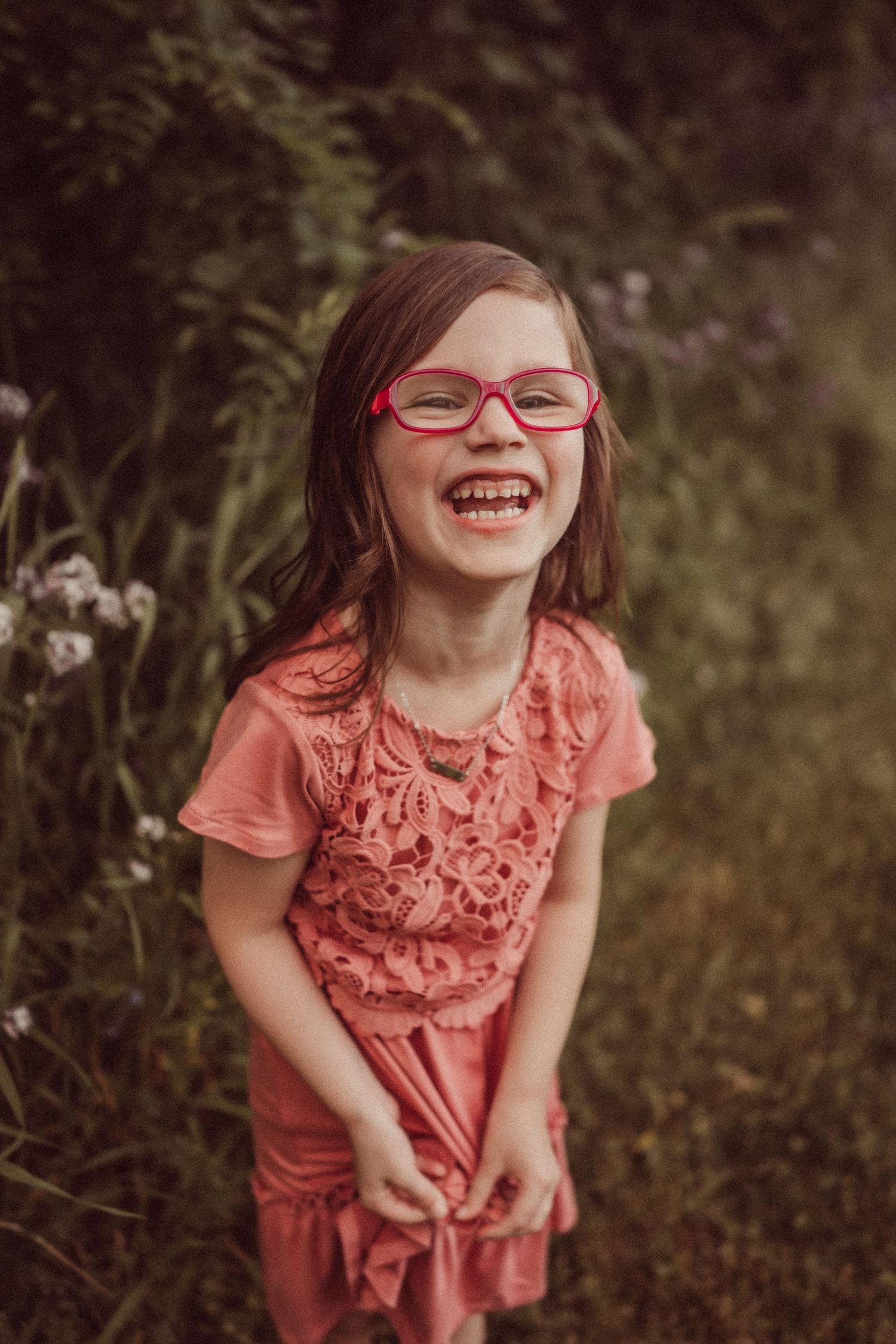 A child in coral colored dress and red glasses laughs joyfully in an outdoor setting.