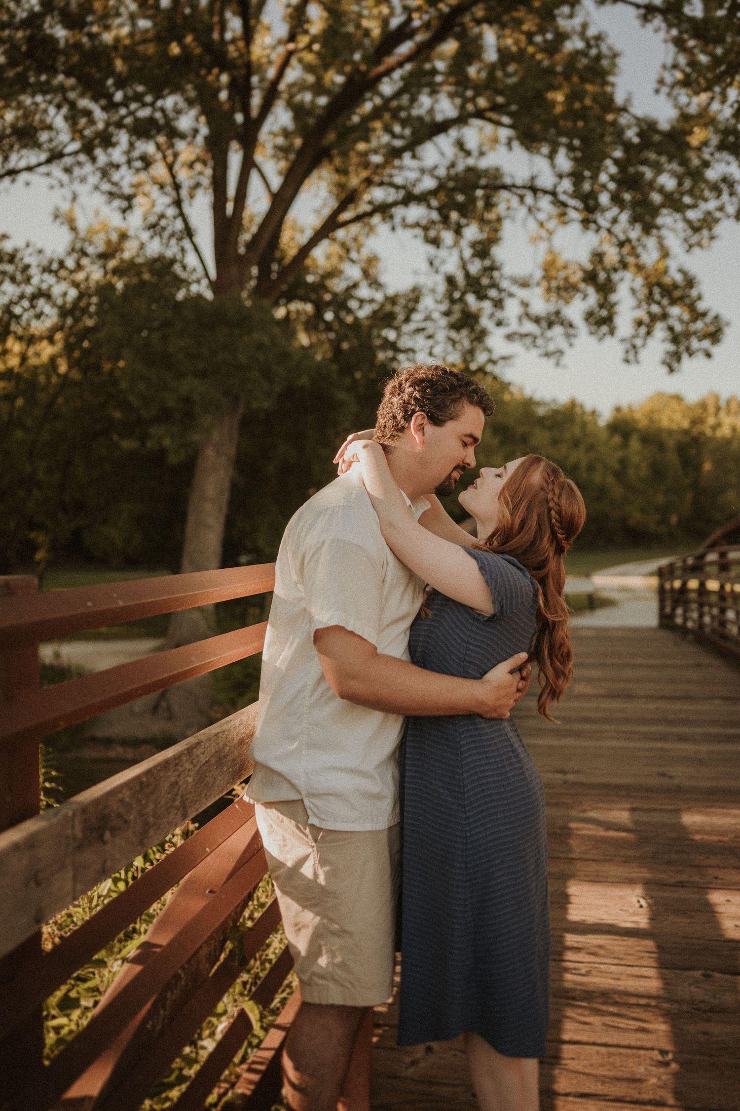 Couple embracing on wooden bridge during golden hour with trees in background.