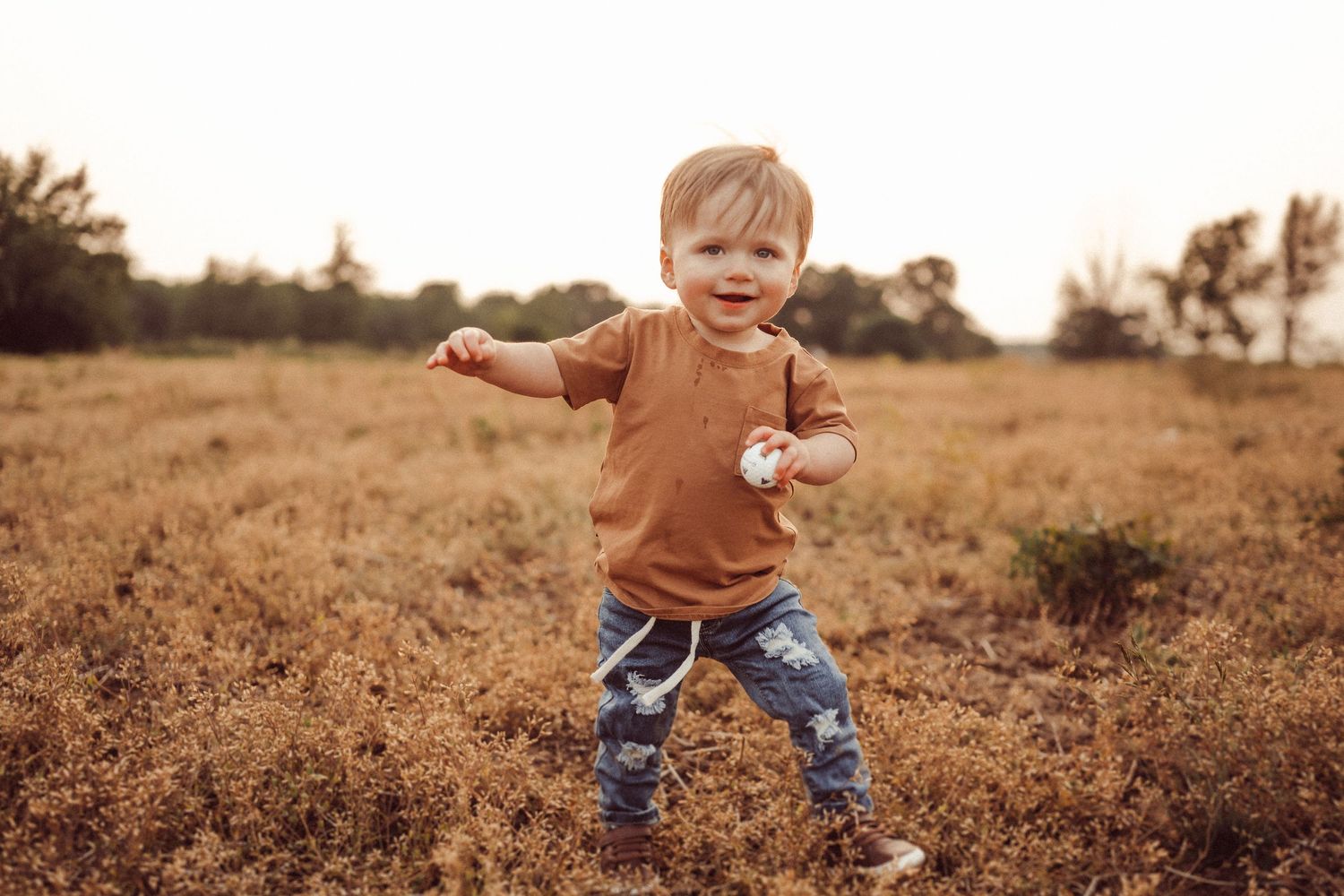 A happy toddler spreads their arms wide while playing in a golden field at sunset.