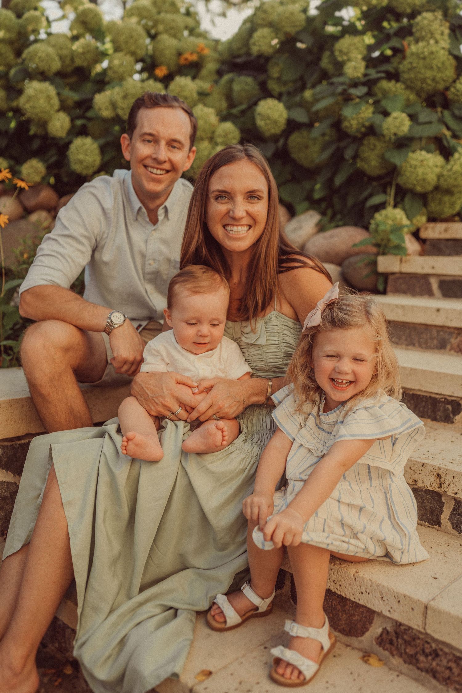 Candid family portrait on stone steps with blooming hydrangeas and orange flowers in background.