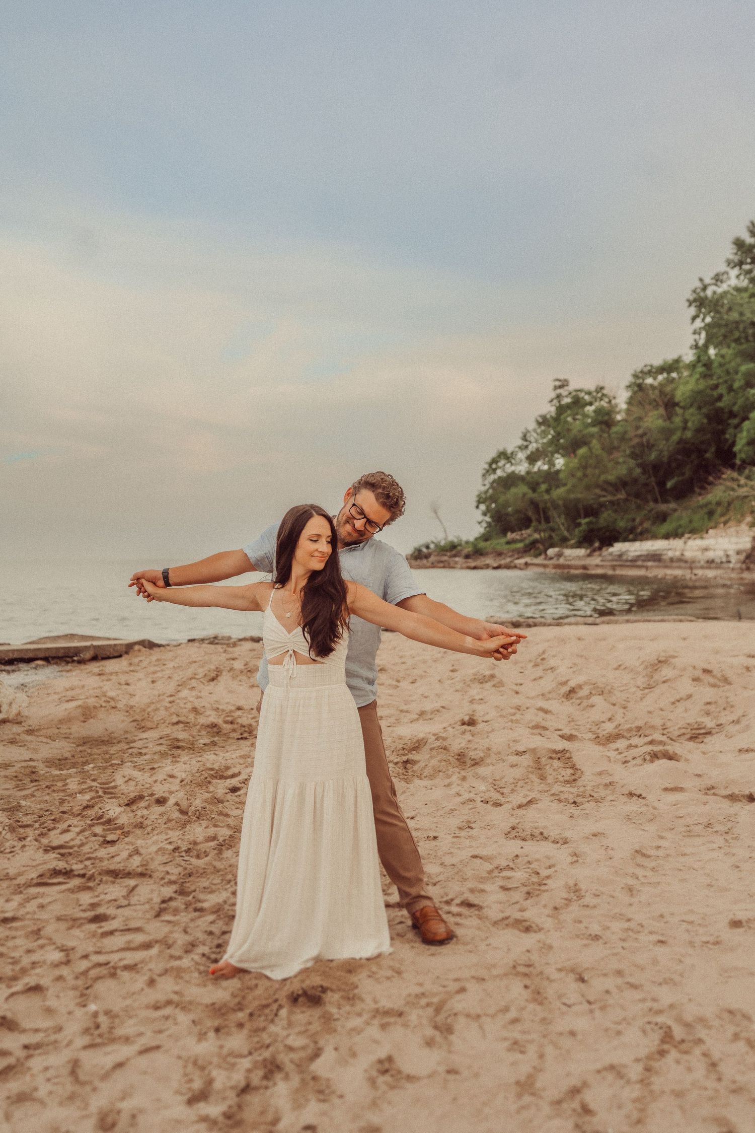Playful couple poses with arms spread wide on sandy beach at sunset.