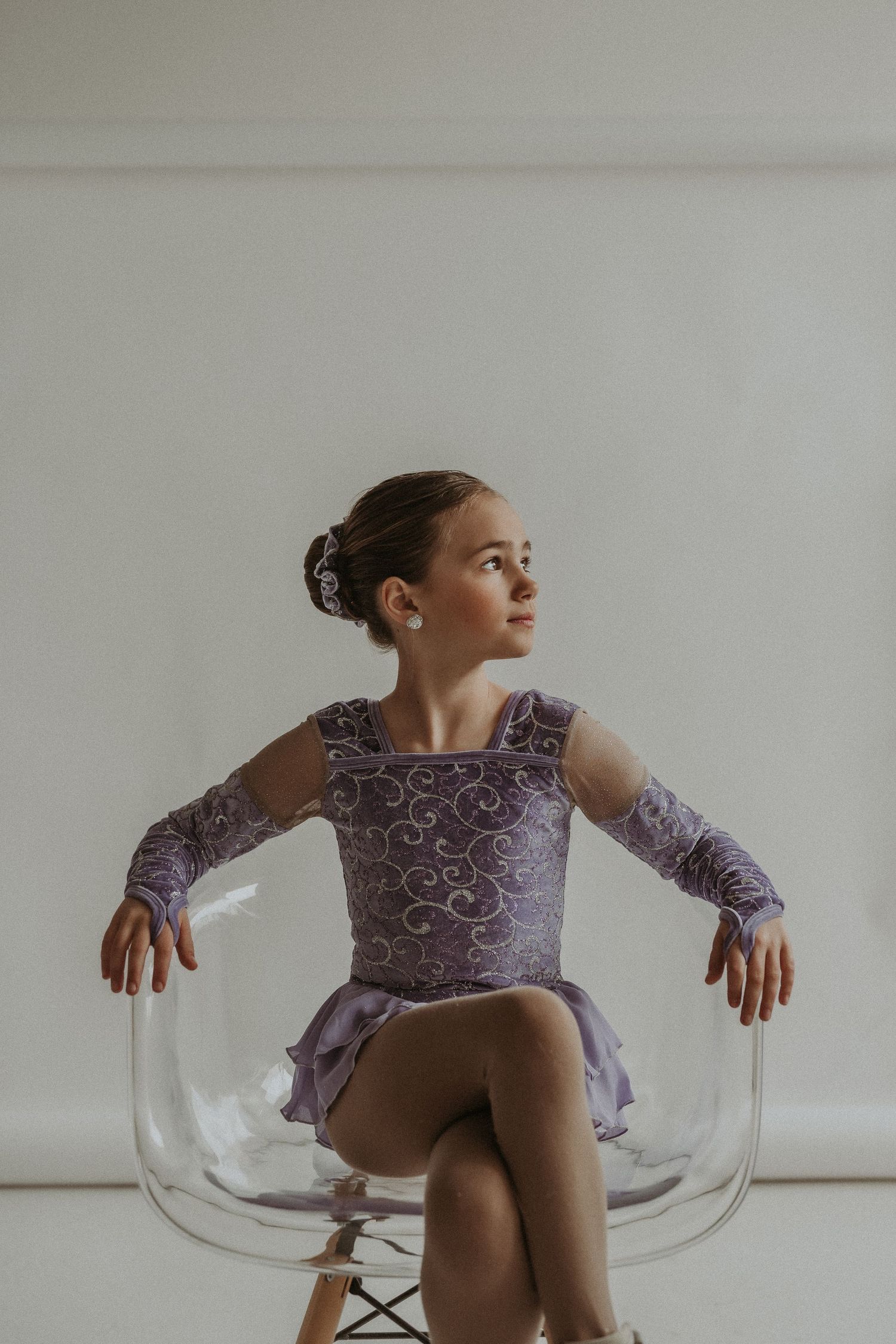 Ice skater in purple lace costume poses gracefully on transparent chair against white backdrop.
