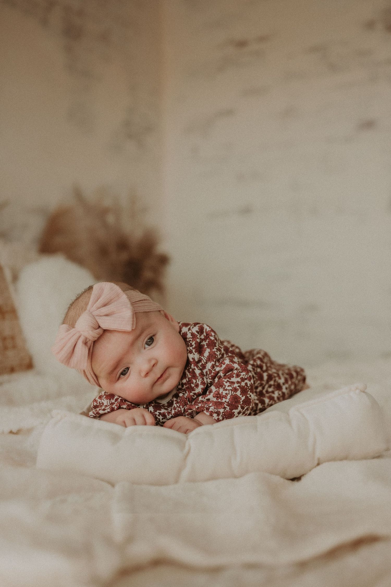 Baby in floral dress and pink bow lounging on cream colored bedding.