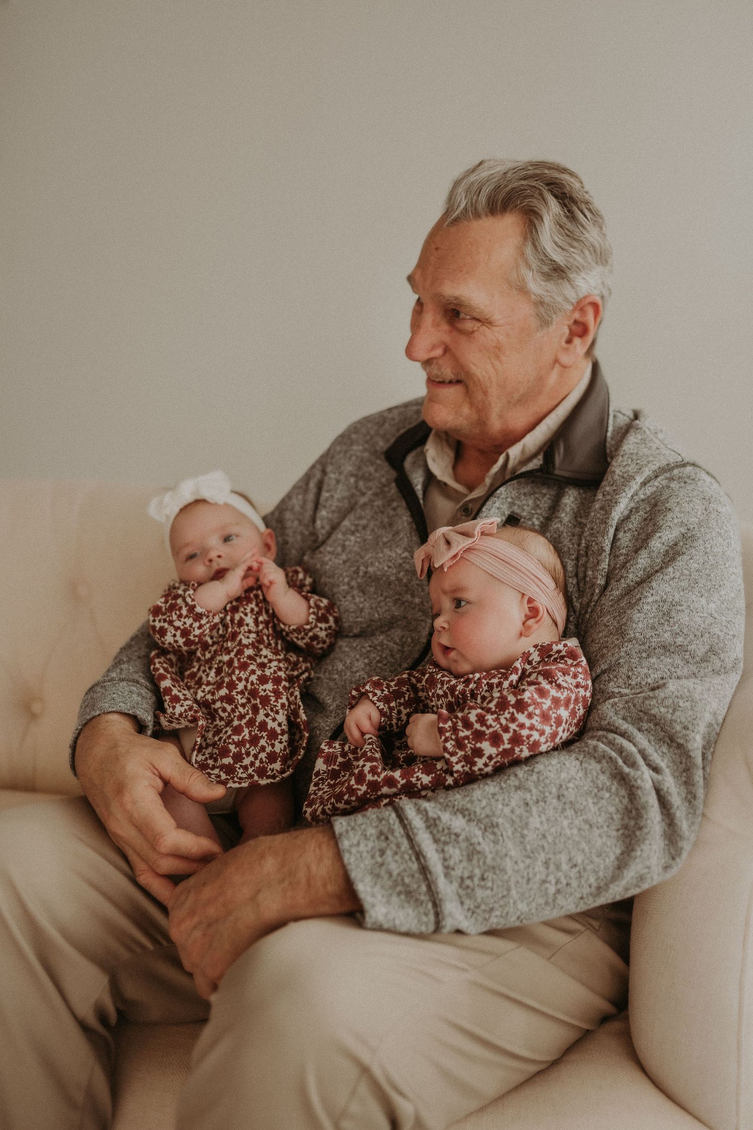 Person in grey sweater shares a tender moment with twin babies in floral outfits on cream couch.
