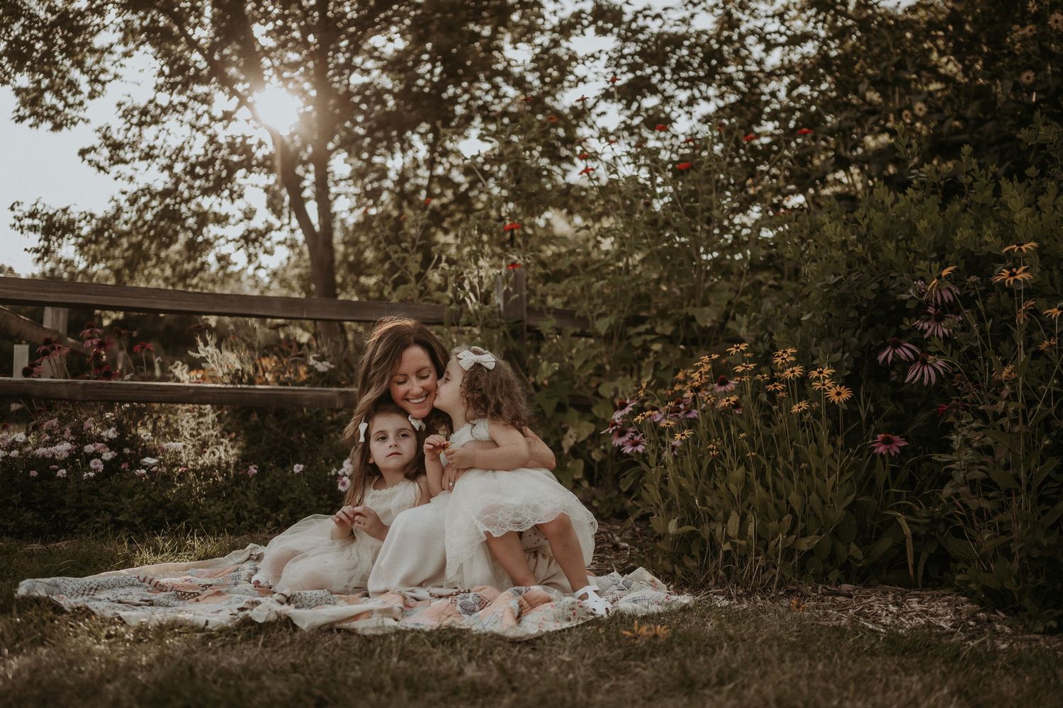 A dreamy outdoor family portrait at sunset with white dresses and a blanket on the grass in a garden setting.
