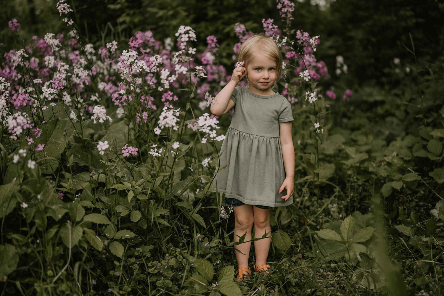 Child in gray dress standing among pink and white flowers in garden setting.