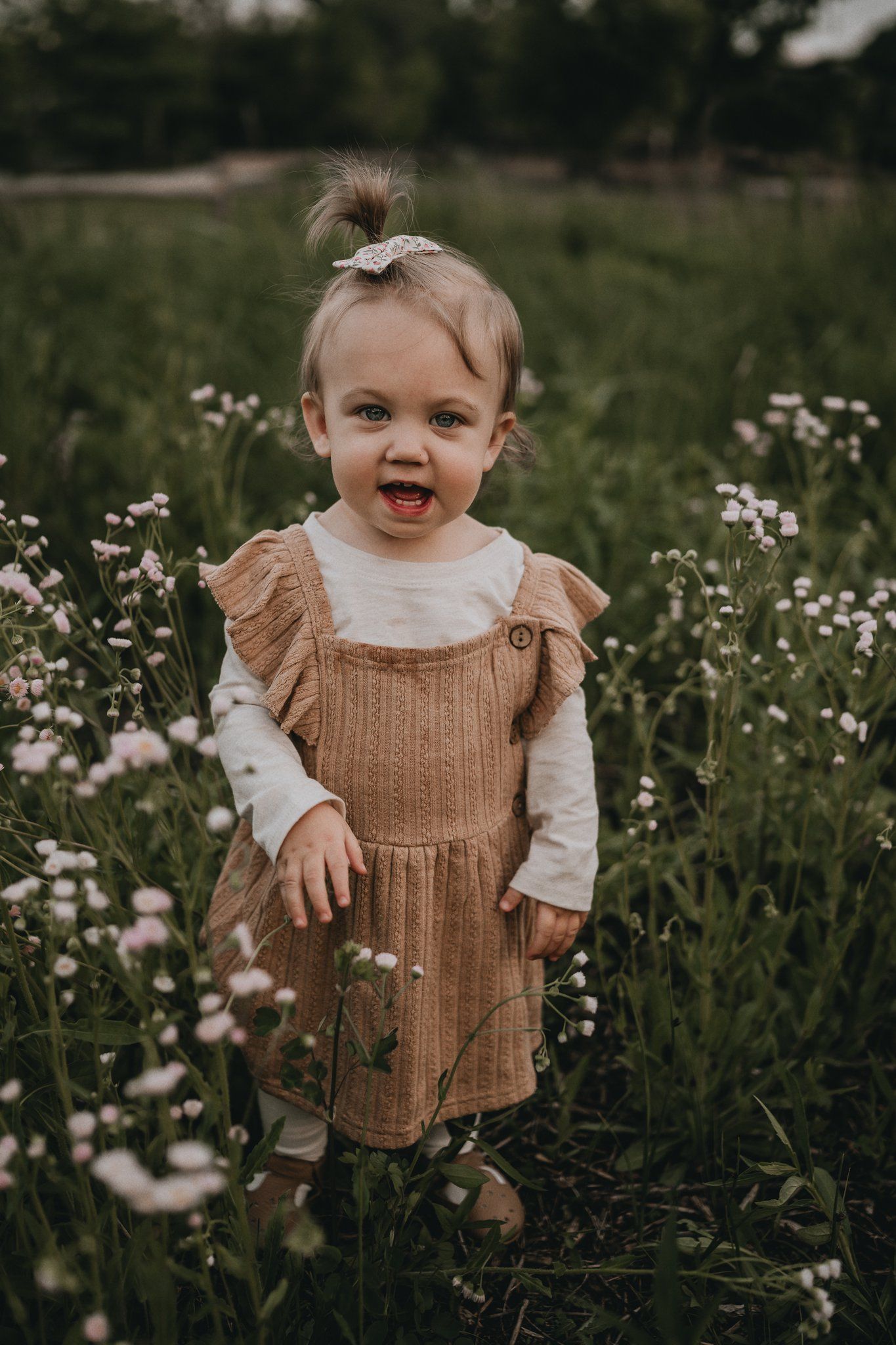 A small child stands joyfully in a field of white wildflowers wearing a brown corduroy dress and white shirt.