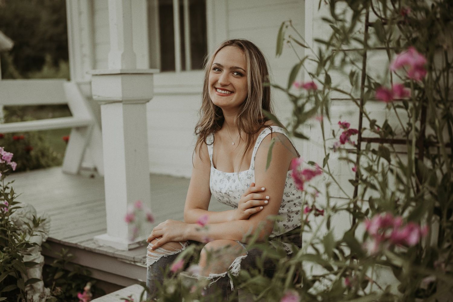 Woman in white top smiles while sitting on porch with pink flowers in background.