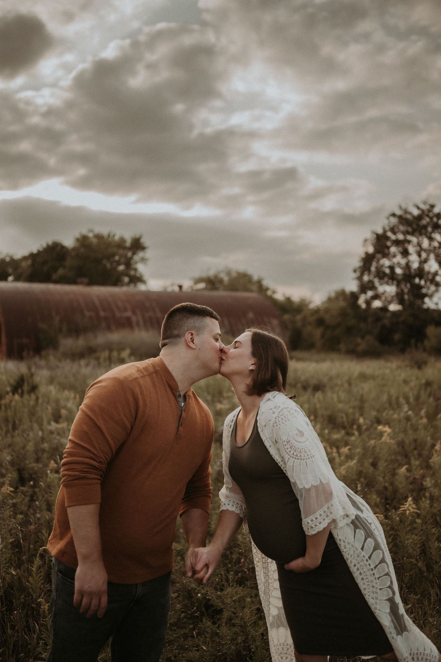 Sweet moment between expecting couple sharing a kiss in front of rustic barn.