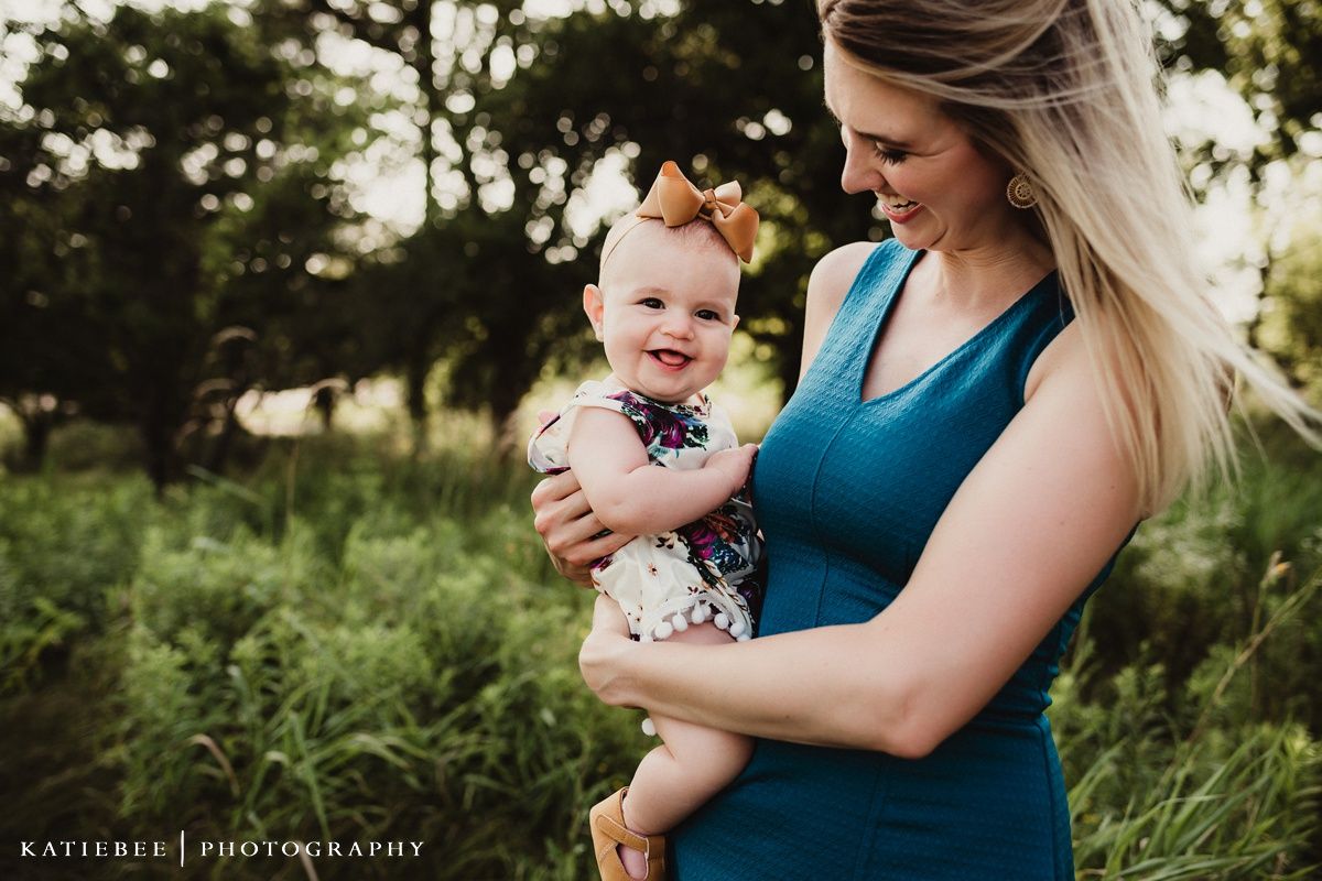 A heartwarming outdoor portrait of a parent in teal dress holding a smiling baby wearing a bow and floral outfit.