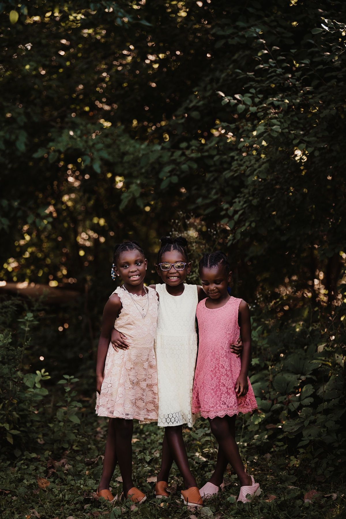 Three children in coordinating pink and white dresses pose together against forest backdrop.