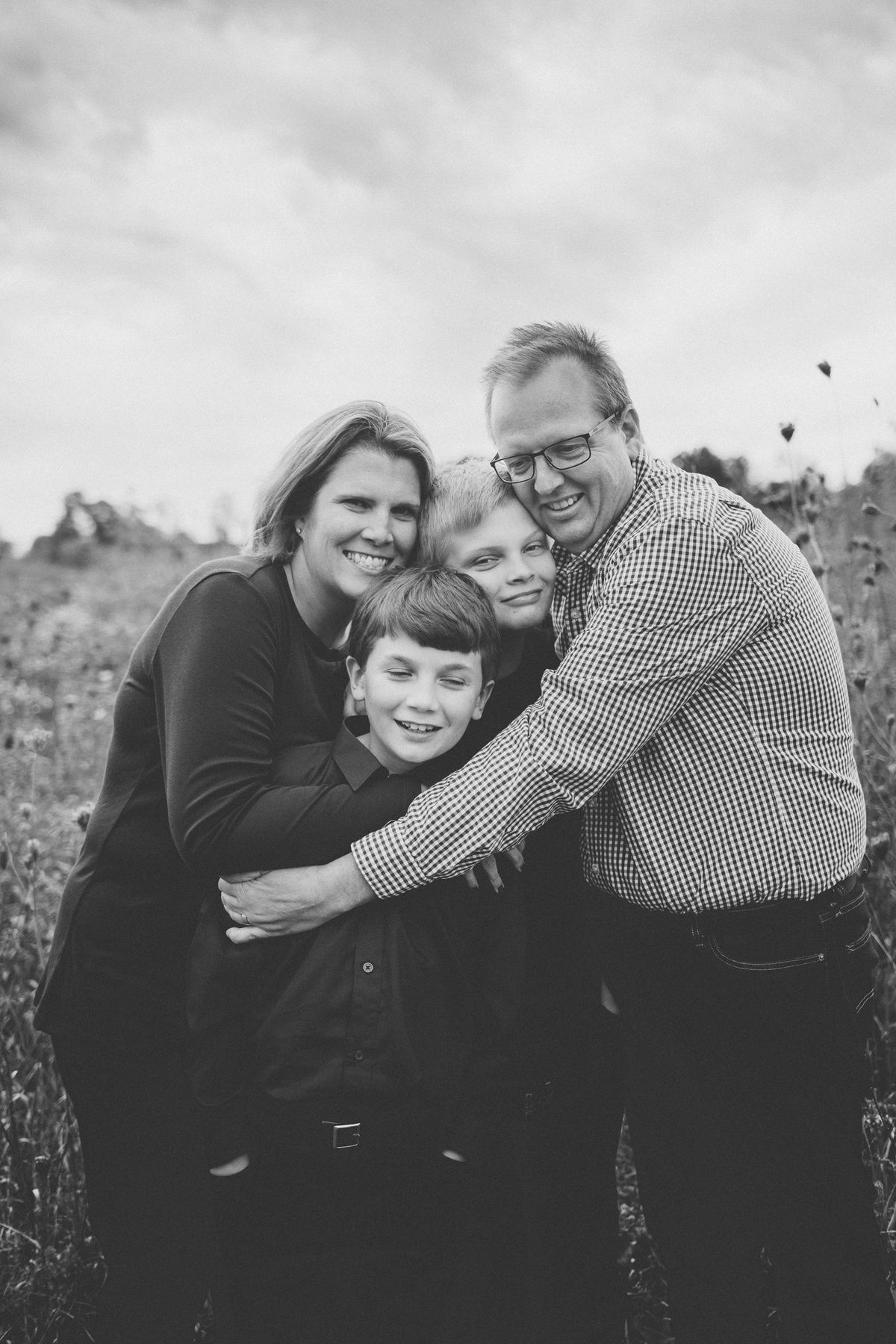 A black and white family portrait shows four people hugging and smiling in a meadow.