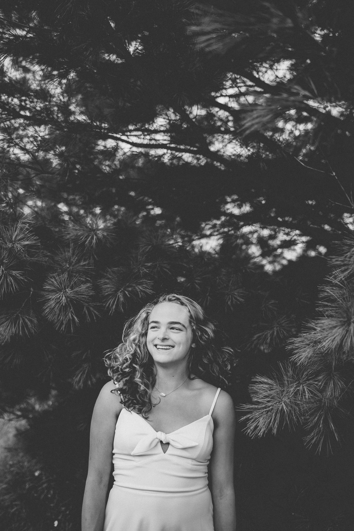 Black and white portrait of someone in a white dress smiling against evergreen branches.