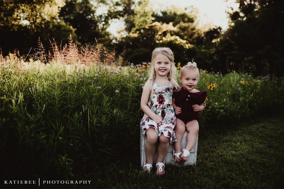 Two children in coordinated outfits sit together on a blanket in a sunlit meadow.