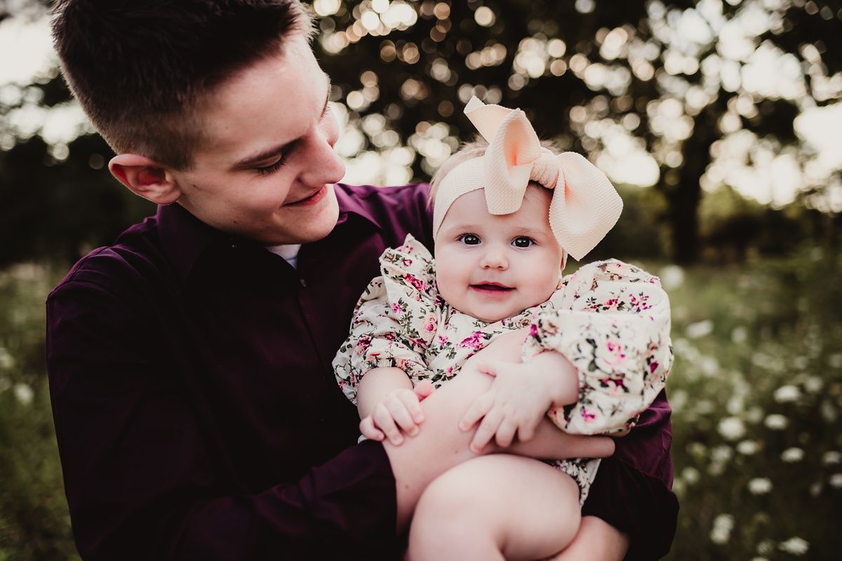 Parent holding smiling baby wearing floral dress and bow against backdrop of white wildflowers.