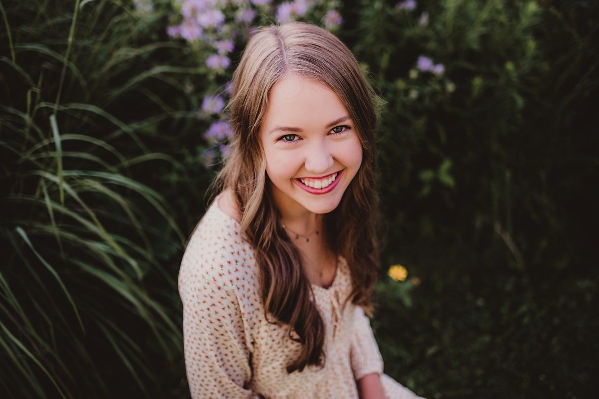 Woman in beige sweater smiling while sitting in garden with purple flowers.