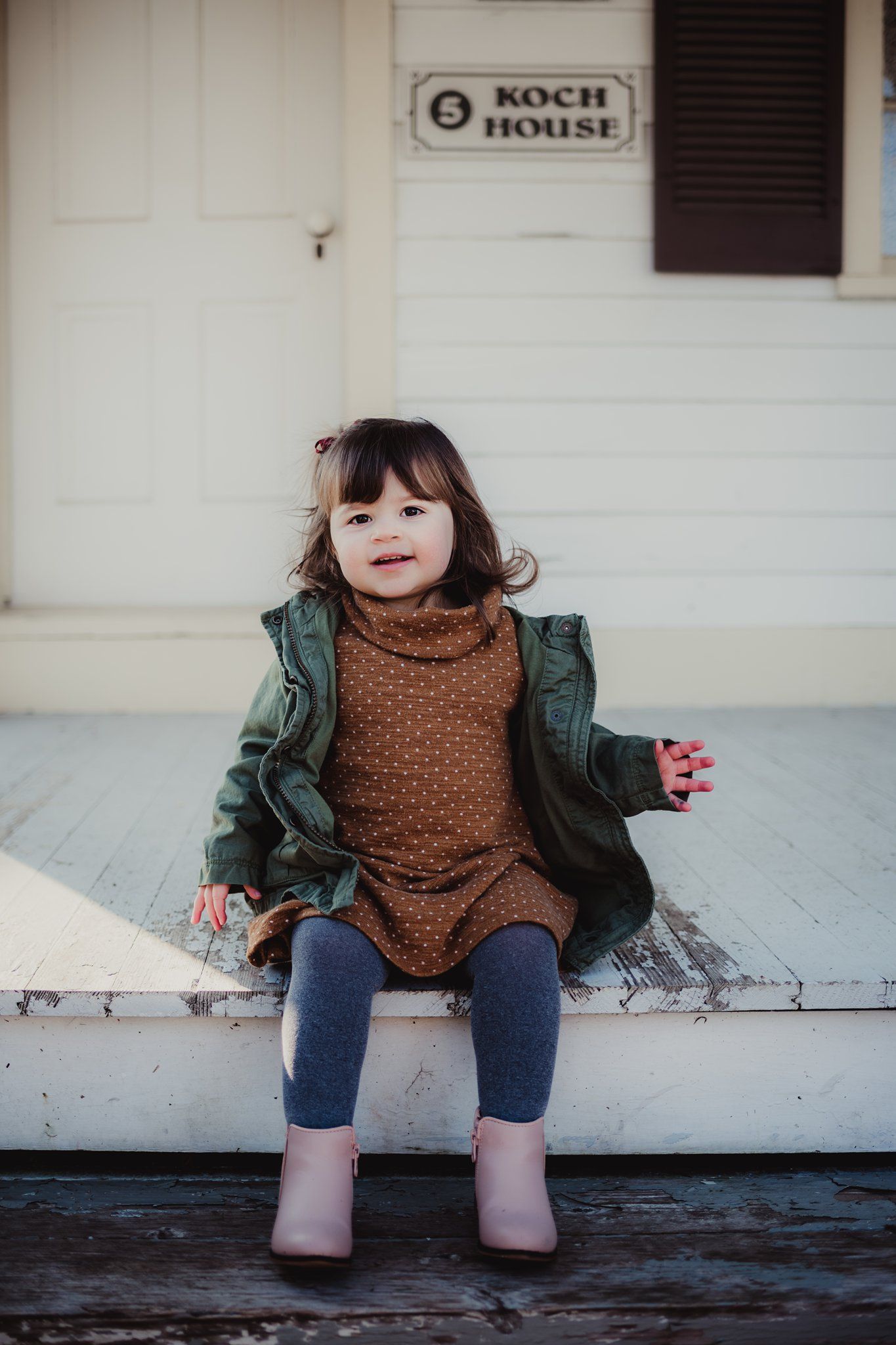 A child sits on wooden steps outside the Koch House wearing a brown dress and green jacket.