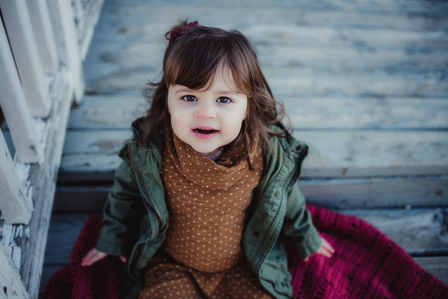 A young child sits on wooden steps wearing a green jacket and brown polka dot dress.