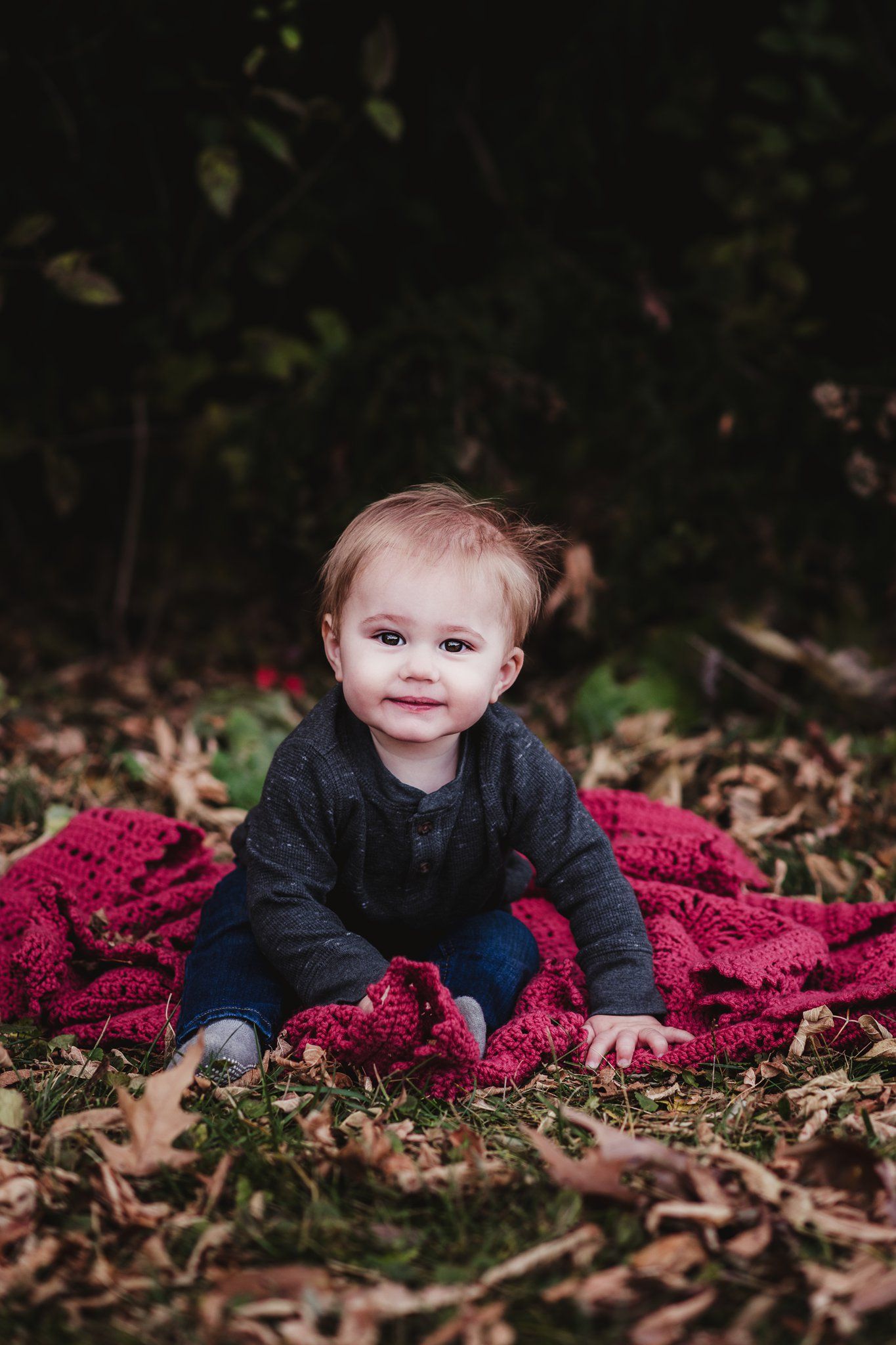 Baby sitting on burgundy blanket among autumn leaves.