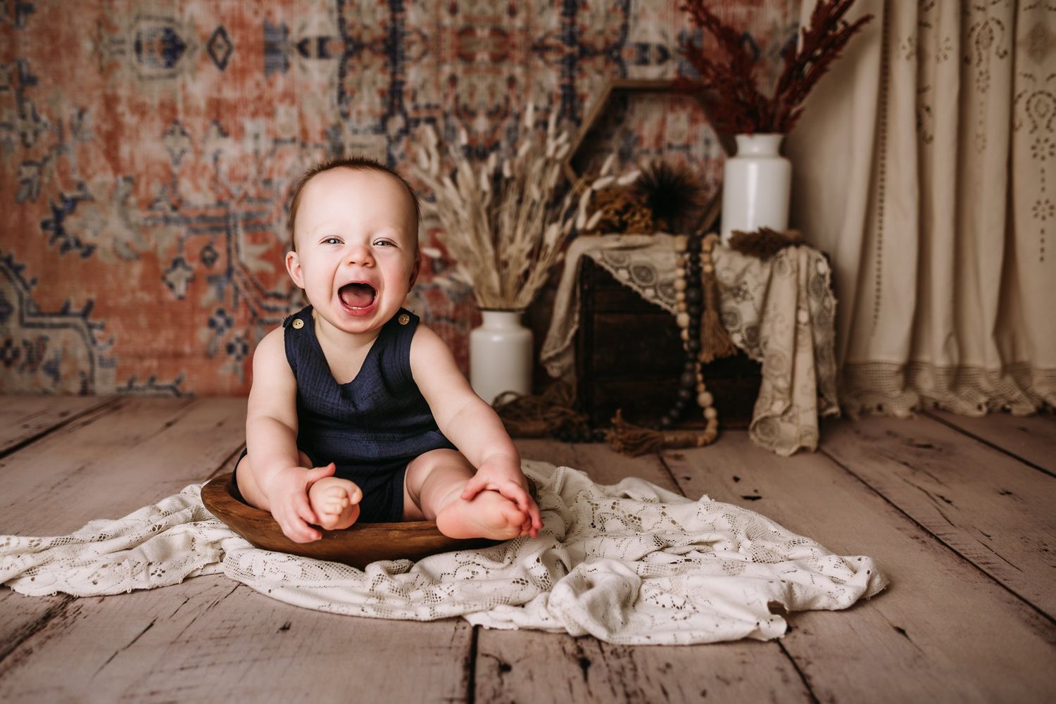 Laughing baby in navy outfit sits in wooden bowl against decorative vintage backdrop.