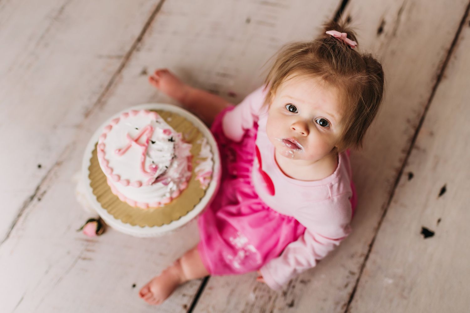Small figure in pink dress sits on wooden floor with decorated celebration cake.