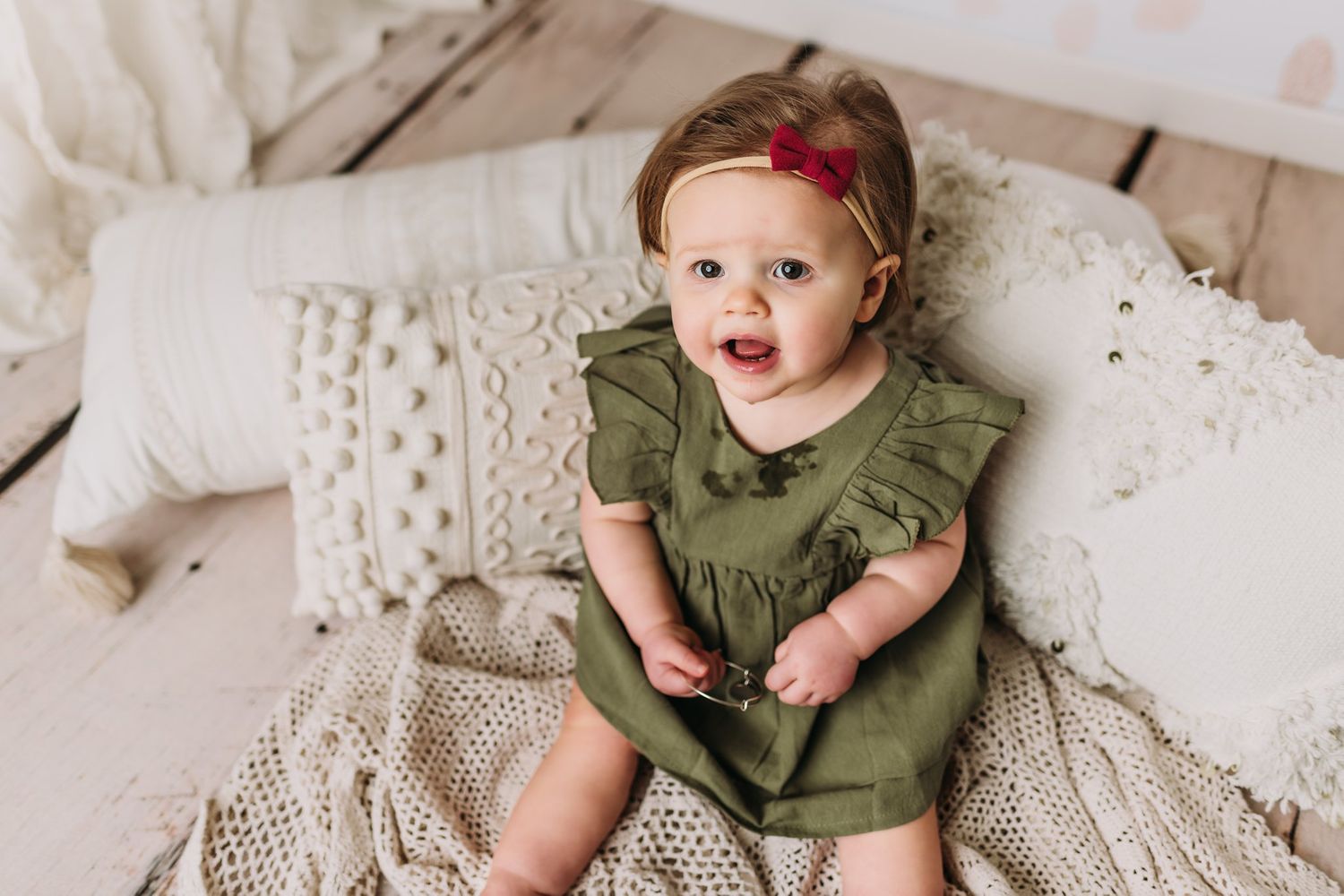 Baby in olive green outfit sits among cream pillows and lace.
