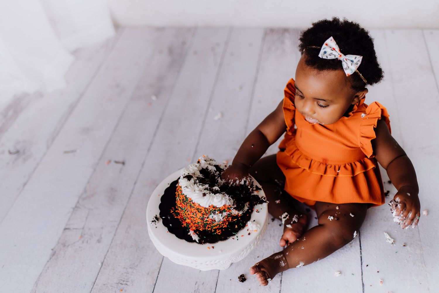 A toddler in orange attire sits beside a messy cake during a cake smash photoshoot.