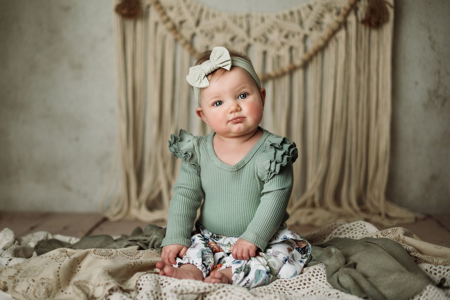 Baby in mint green outfit sits against macrame backdrop on cream textured blanket.