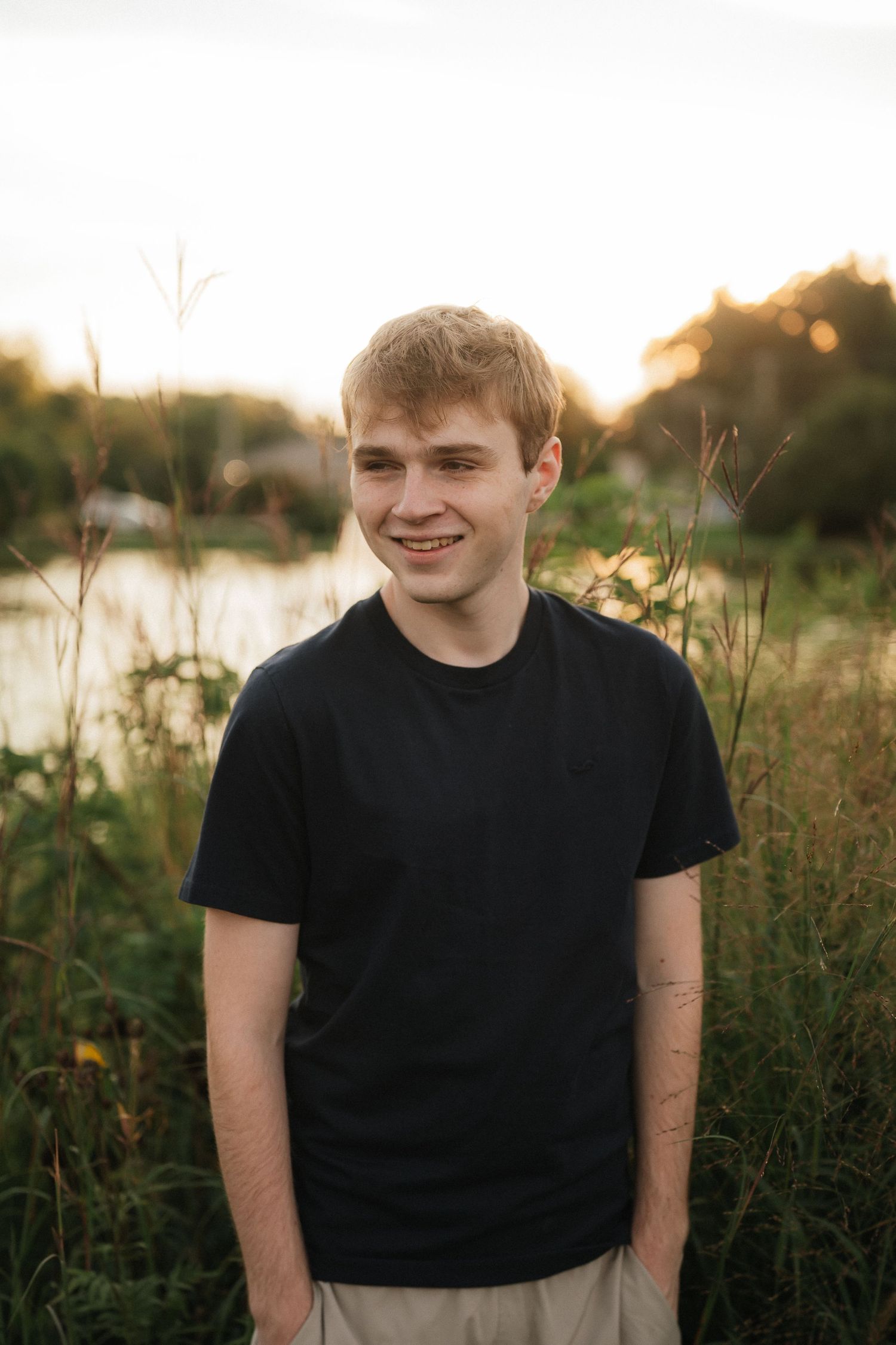 Person in black t-shirt smiling outdoors during golden hour sunset among tall grass.