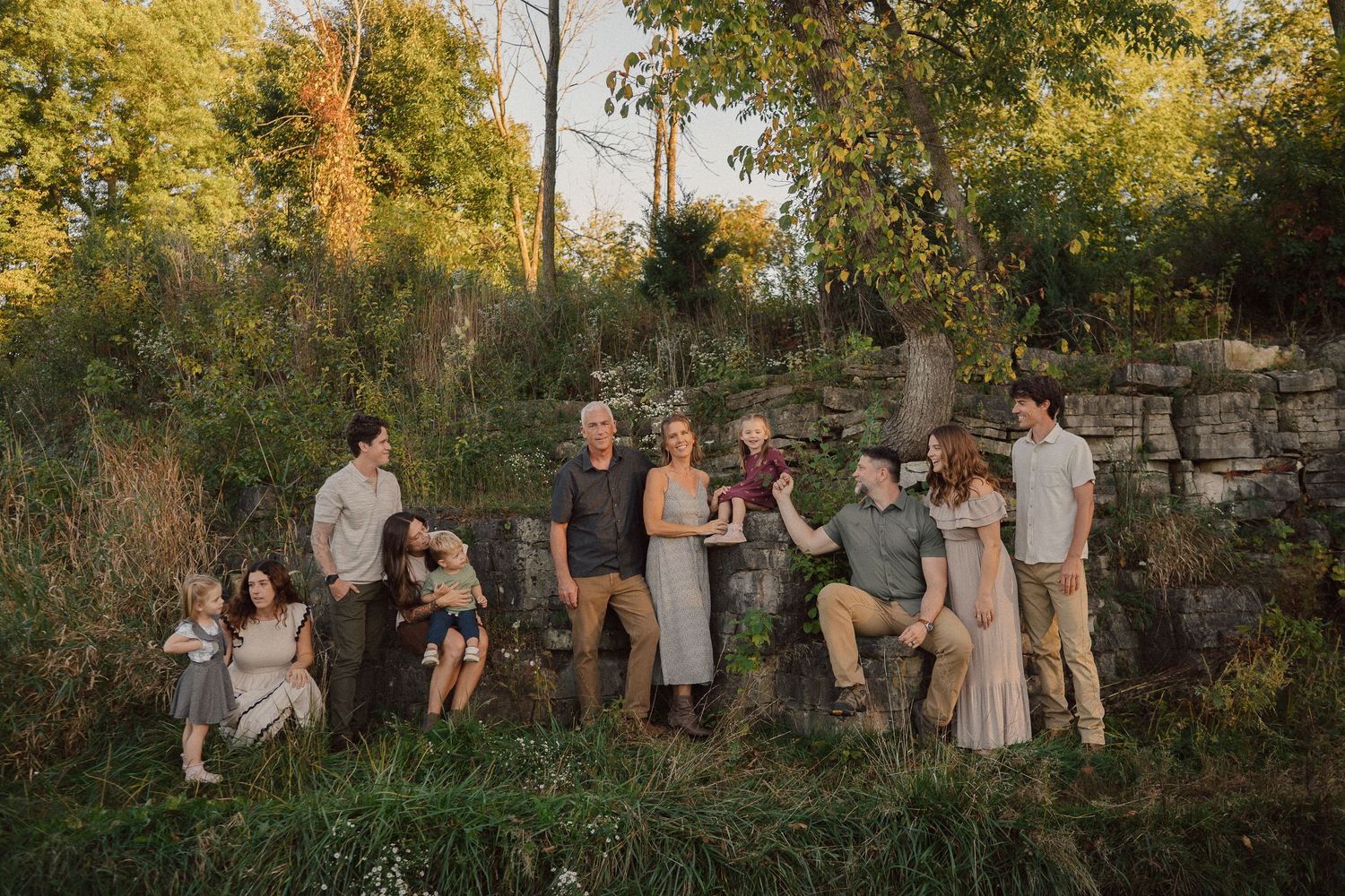 Extended family posing together in natural outdoor setting during sunset with stone wall in background.