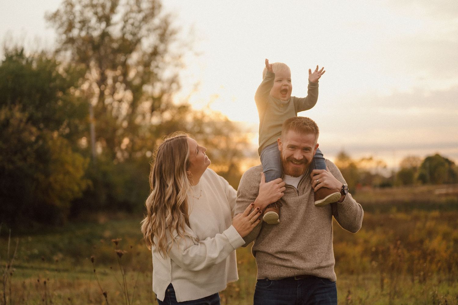 A family plays together in a sunlit meadow during golden hour as their child sits on shoulders.