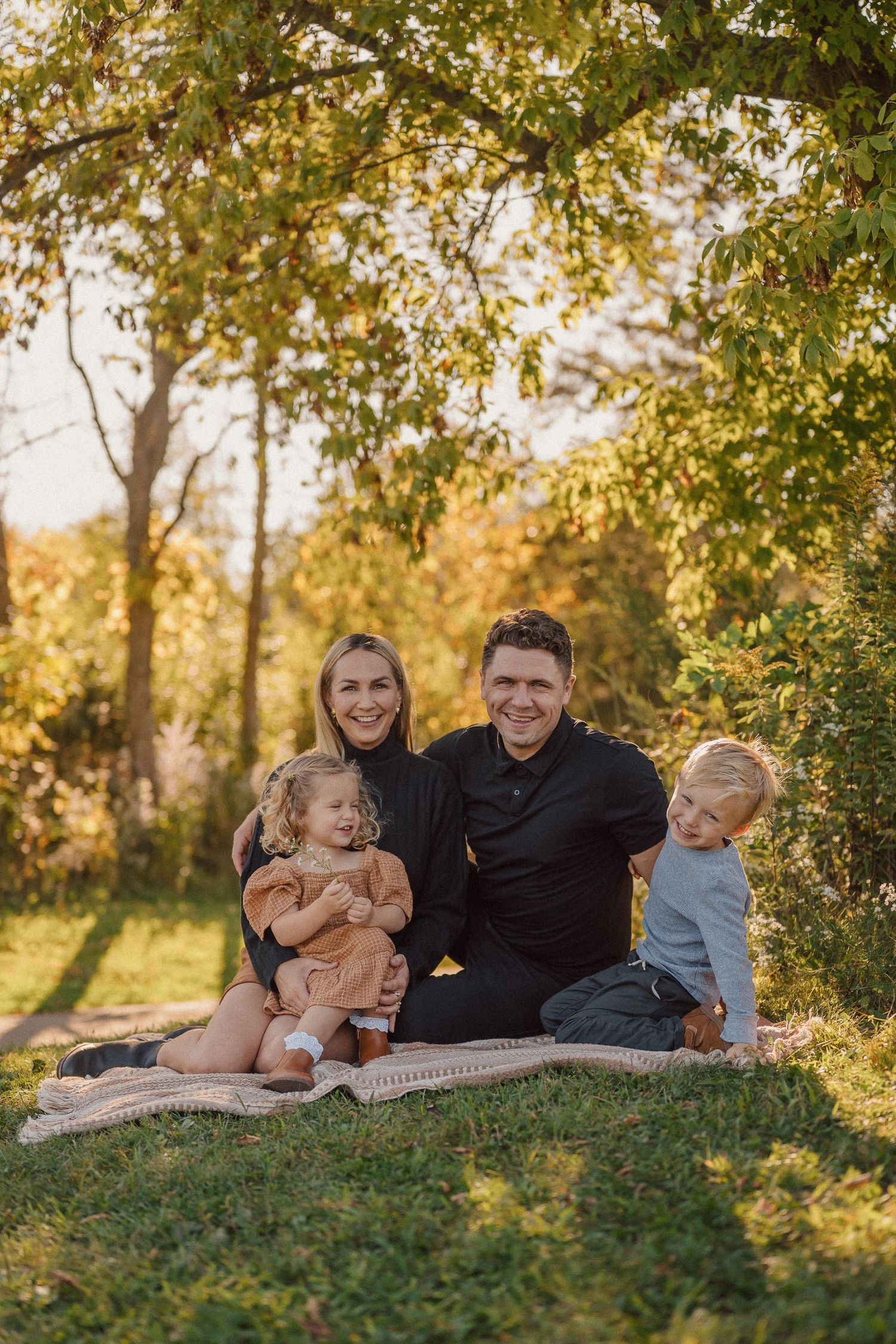 A family relaxes together on a grassy hillside during golden hour with autumn trees in the background.