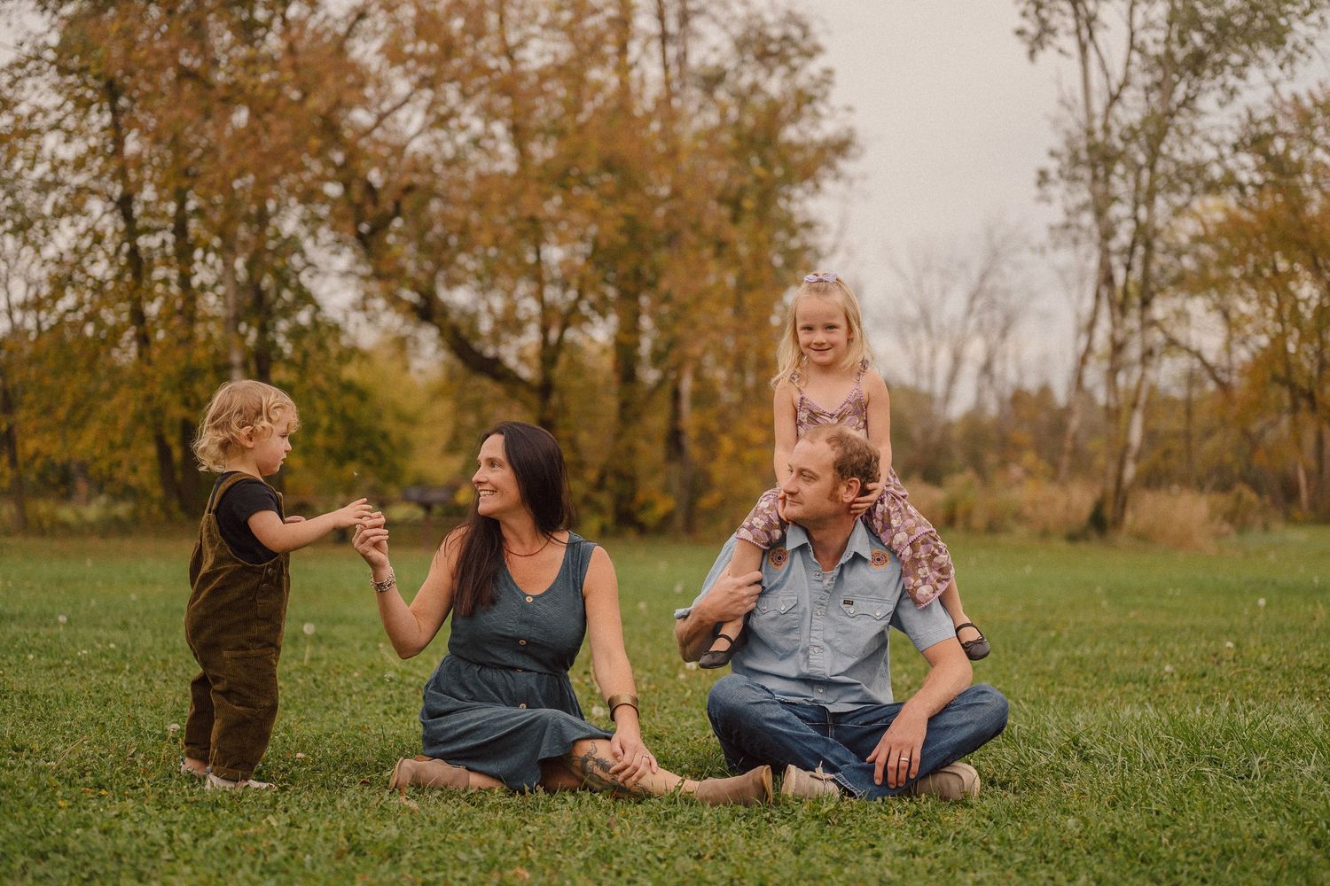 A playful family moment on grass with autumn trees in the background.