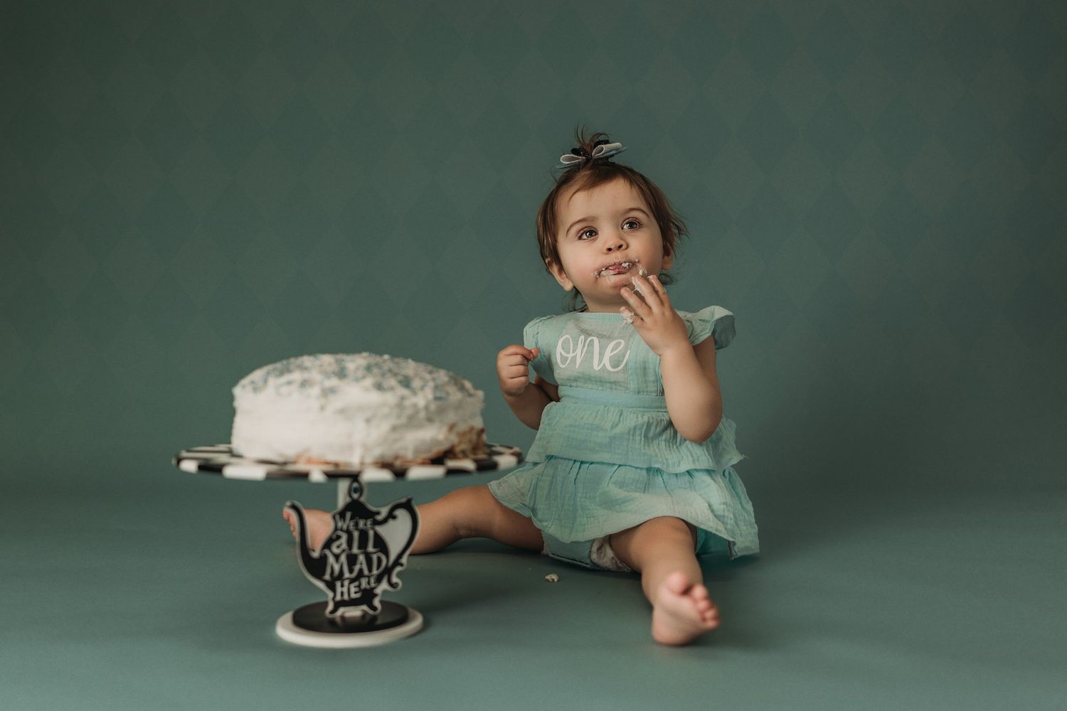 A mint green dress adorns a small subject seated beside an ornate cake stand against teal backdrop.