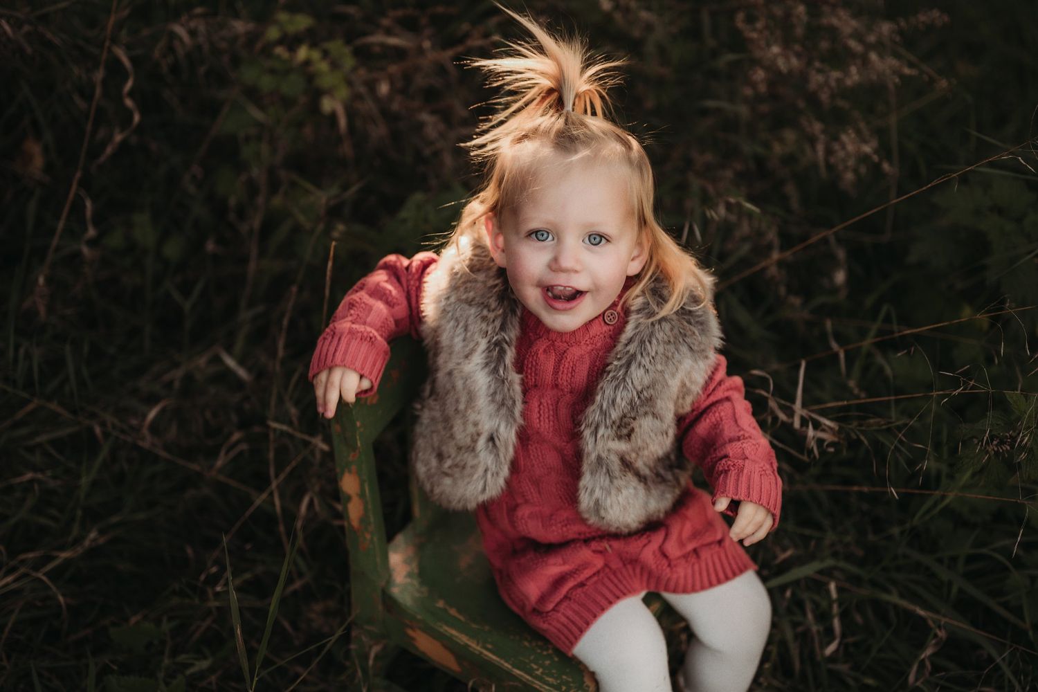 Child in red sweater and fur vest sitting on wooden chair outdoors.