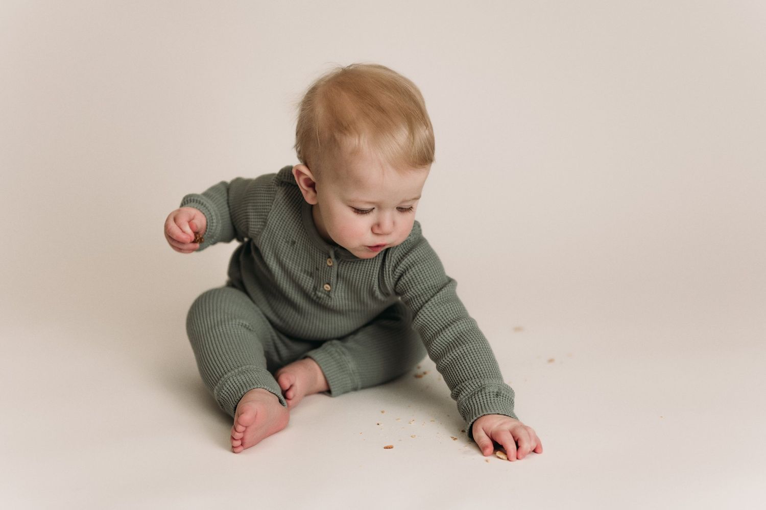 Baby in olive green outfit explores while crawling on neutral background.