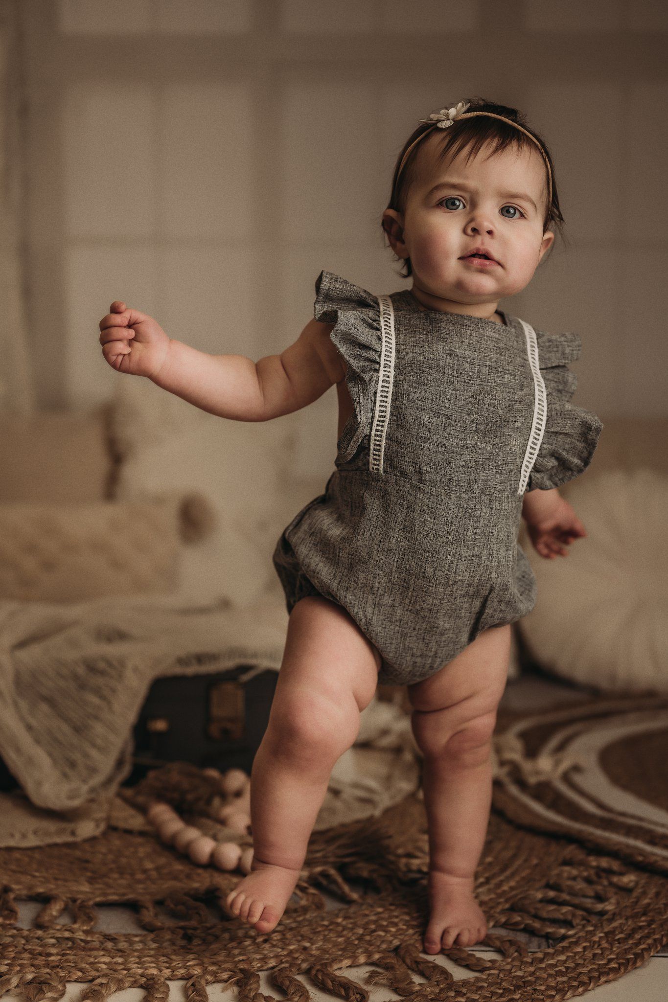 A toddler in a gray ruffled romper standing on a textured rug with vintage decor.