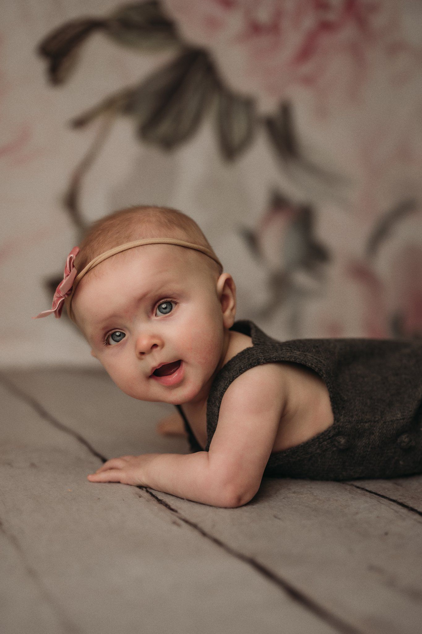Baby in dark outfit lying on floor with pink floral backdrop.