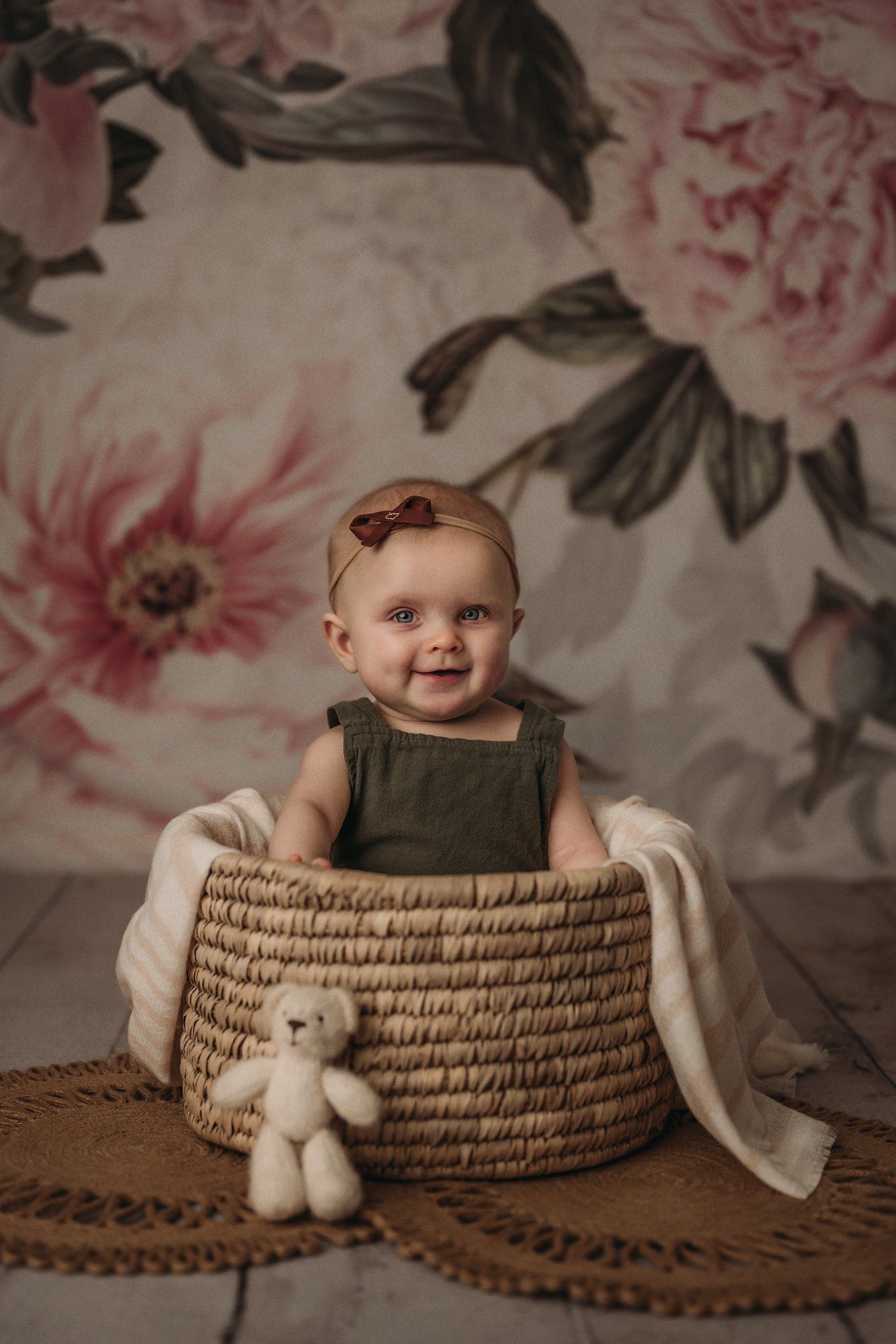 Child sitting in woven basket against floral backdrop with white teddy bear.