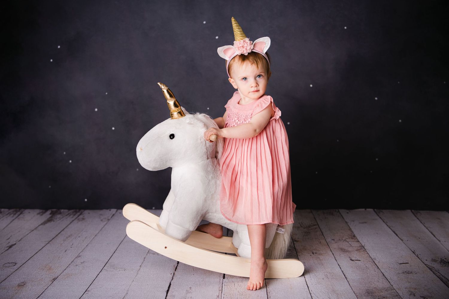 Child in pink dress riding rocking unicorn toy against dark backdrop with sparkles.