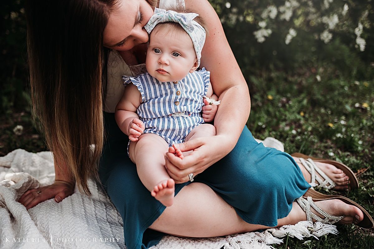 Parent and baby sitting on white blanket outdoors, baby wearing blue striped romper and bow.