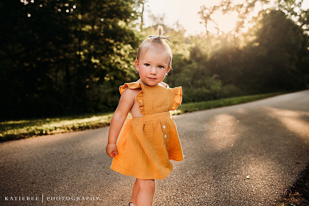 Young child in orange ruffled dress standing on path during sunset.