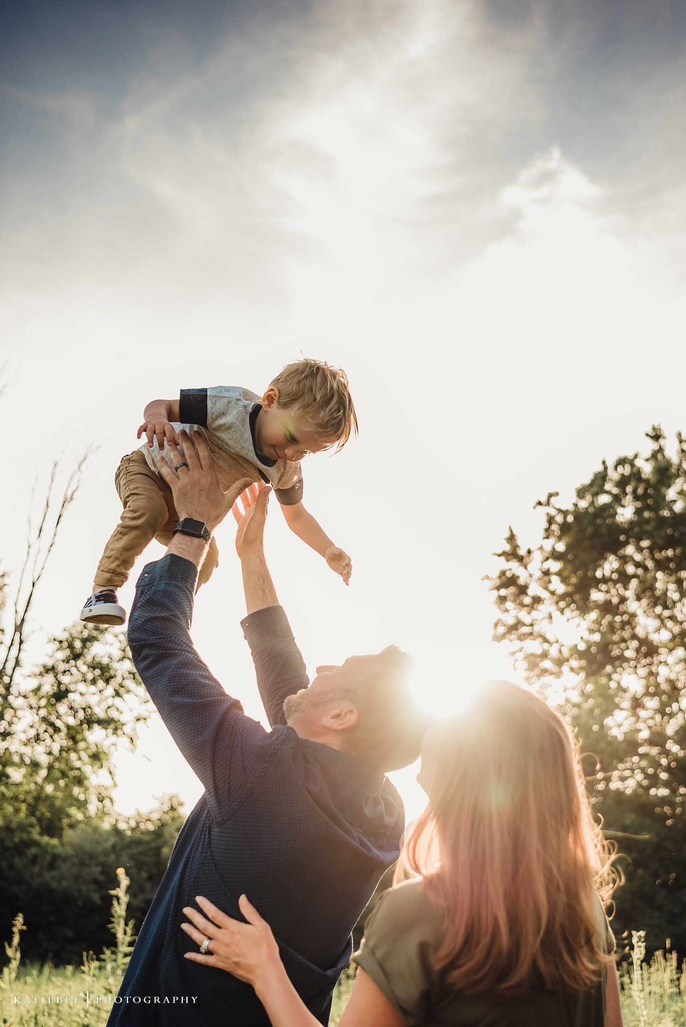 Parents lift their child playfully in the air against a dramatic sunset sky.