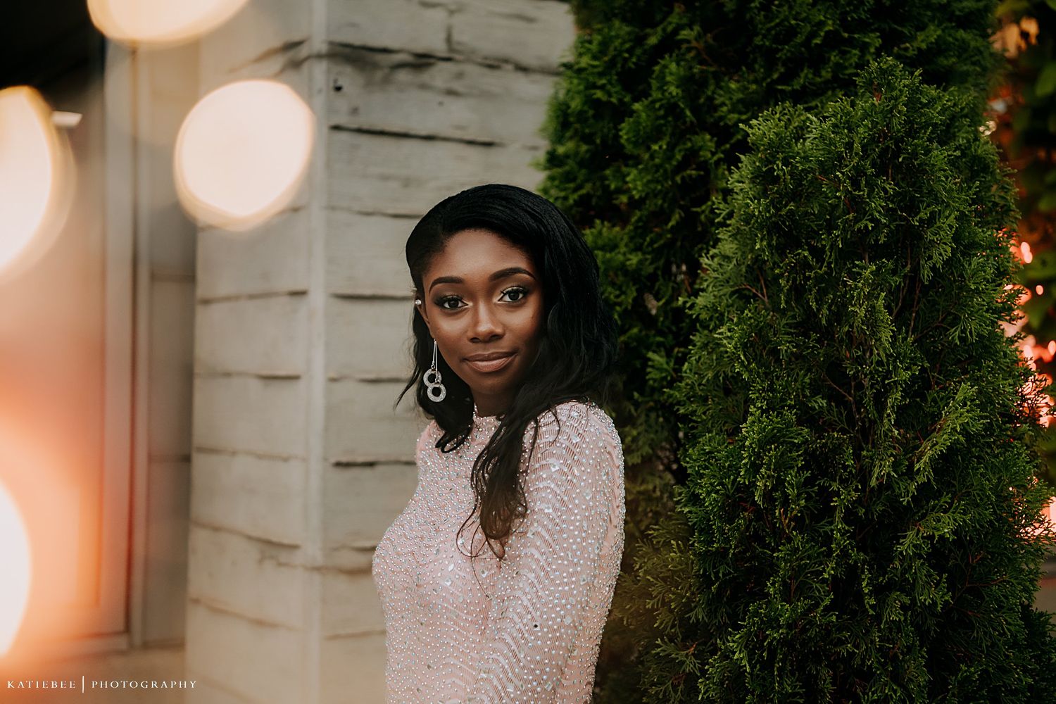 A woman in a sparkling beige dress poses against a white brick wall with greenery and bokeh lights.