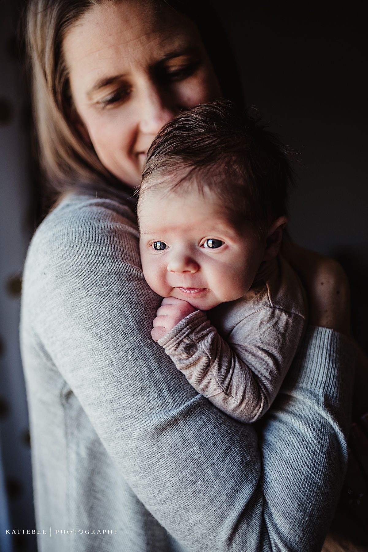 Intimate portrait of caregiver in grey sweater holding infant against dark background.