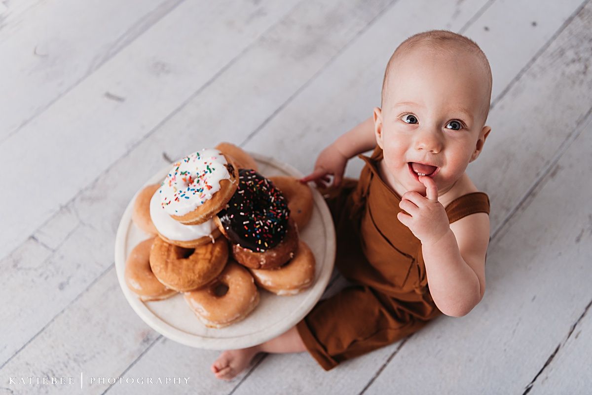 Baby in brown outfit sits on wooden floor next to decorative plate of frosted donuts.